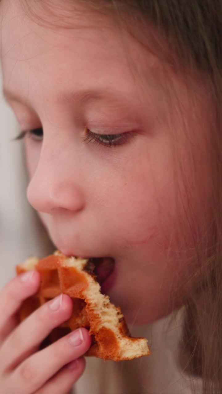 Close up of young girl eating waffle indoors with soft lighting. Hand holding partially eaten piece while focused on act of consuming food. Background blurred emphasizing subject