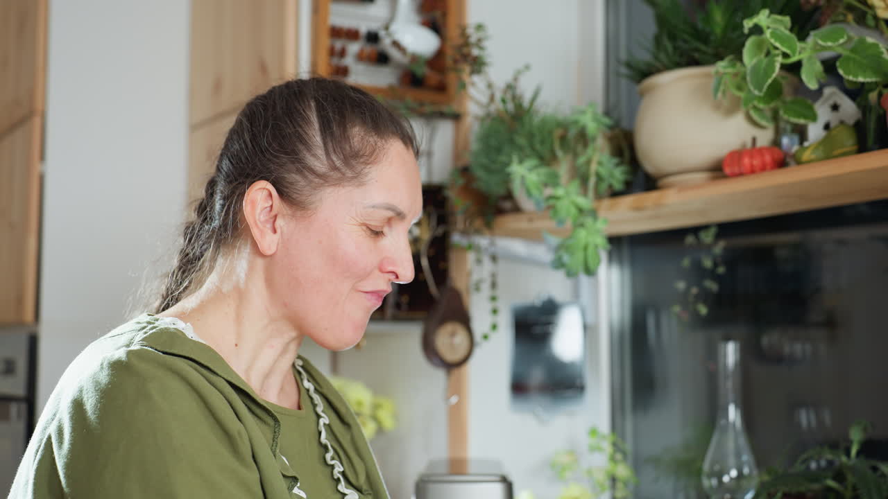 White woman in green dress eats fresh berry in cozy kitchen, smiling and shaking head joyfully as she enjoys taste, surrounded by indoor plants and natural light
