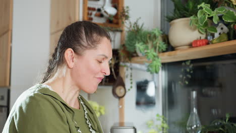 white woman in green dress eats fresh berry in cozy kitchen, smiling and shaking head joyfully as she enjoys taste, surrounded by indoor plants and natural light