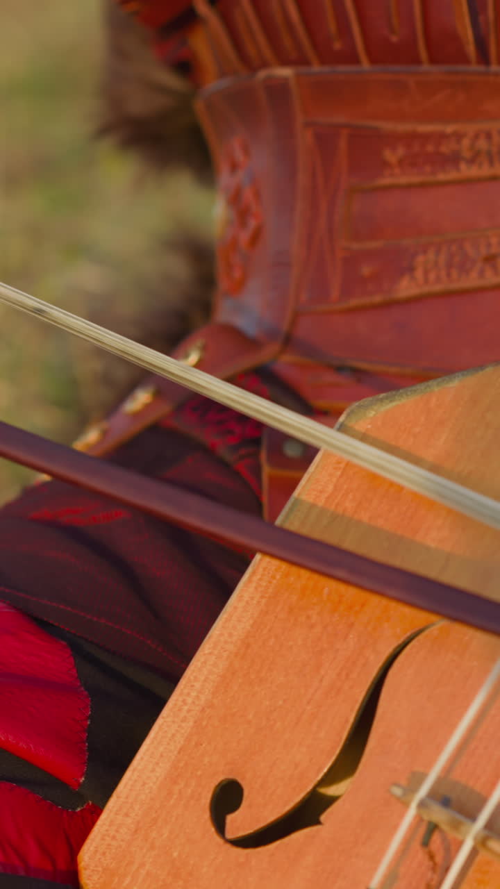 Musician in ethnic leather clothes plays igil fiddle-bow instrument sitting on green field closeup slow motion. Representation of old Yakut music