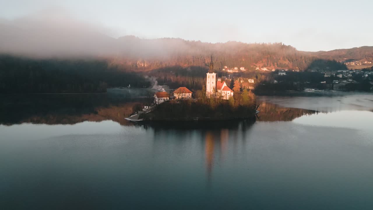 In una fredda mattina d'inverno con una magnifica alba, il filmato aereo si eleva e mostra l'iconica isola con la chiesa nel lago di Bled, Slovenia, mentre nuvole nebbiose fluttuano e si riflettono lungo il lago.