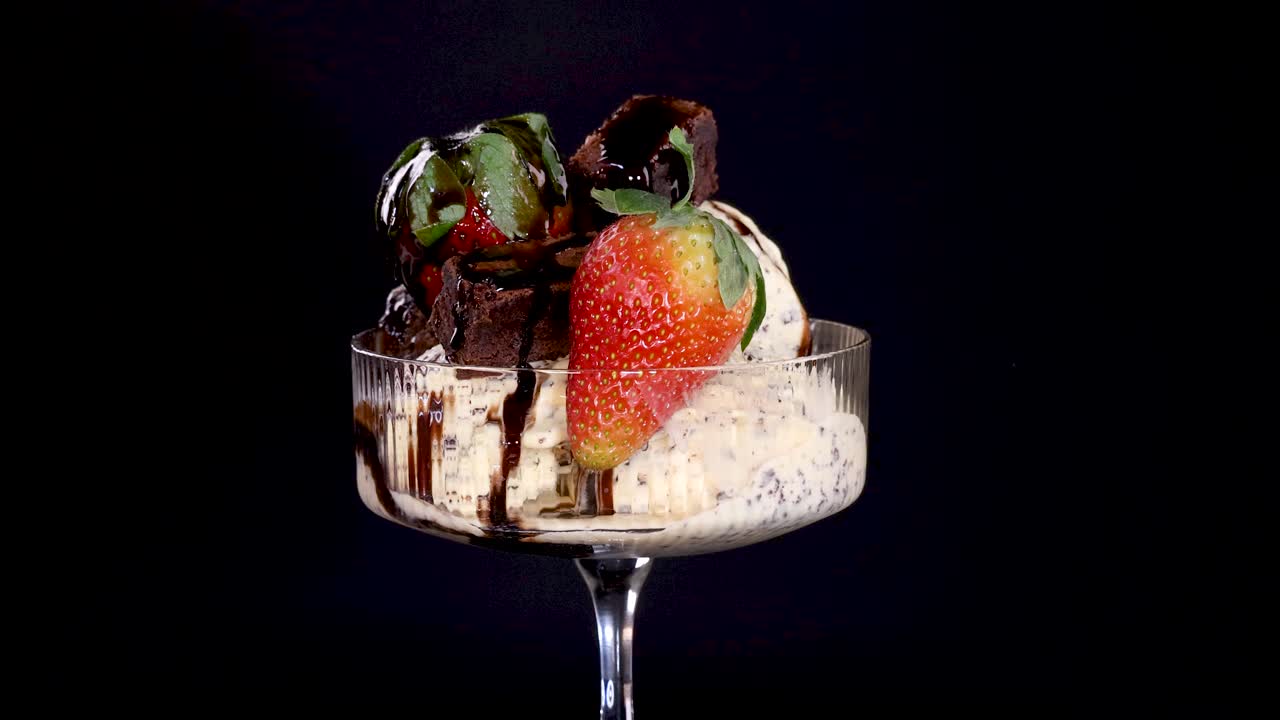 A hand places a large strawberry onto a glass dish of vanilla ice cream, chocolate sauce, brownie pieces, and chocolate chips against a black background with studio lighting