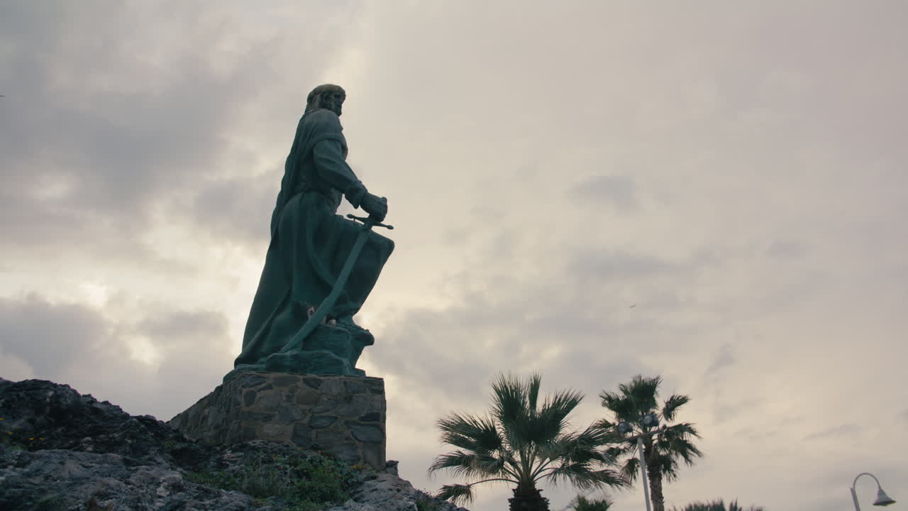 Vista dal basso di una statua storica che impugna una spada, eretta su un piedistallo di pietra sopra un terreno roccioso con palme e nuvole drammatiche. Un gatto randagio vi si riposa sopra