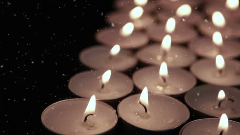 animation of white snowflakes falling over lit tea light candles on black background