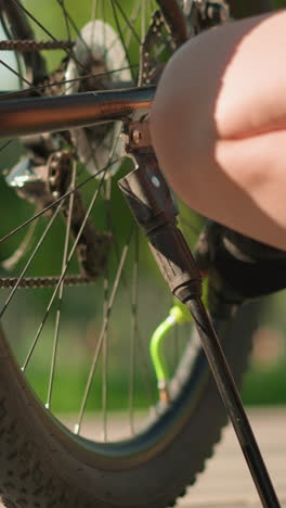 low-angle view of woman squatting next to bicycle, pumping air into tire using green nozzle, sunny outdoor setting with blurred background of fence, trees, and passerby