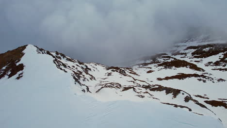 Vista-De-Pájaro-De-Pedestal-Ascendente-Aéreo-De-Una-Persona-Tendida-En-Un-Suelo-Cubierto-De-Nieve-Y-Luego-Revelando-Un-Acantilado-Montañoso-Empinado-Sobre-Las-Nubes-Y-Panoramizando-Hasta-Una-Brillante-Puesta-De-Sol