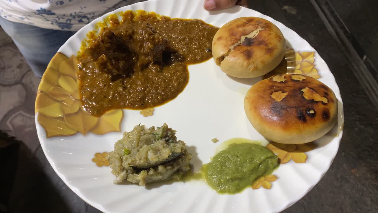 en un video se puede ver a un hombre sosteniendo un plato de sabroso litti chokha empapado en ghee y servido con berenjena asada o baingan bharta, chutney picante verde y curry de cordero kasa