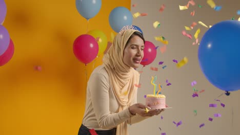 studio portrait of woman wearing hijab and birthday headband celebrating birthday blowing out candles on cake