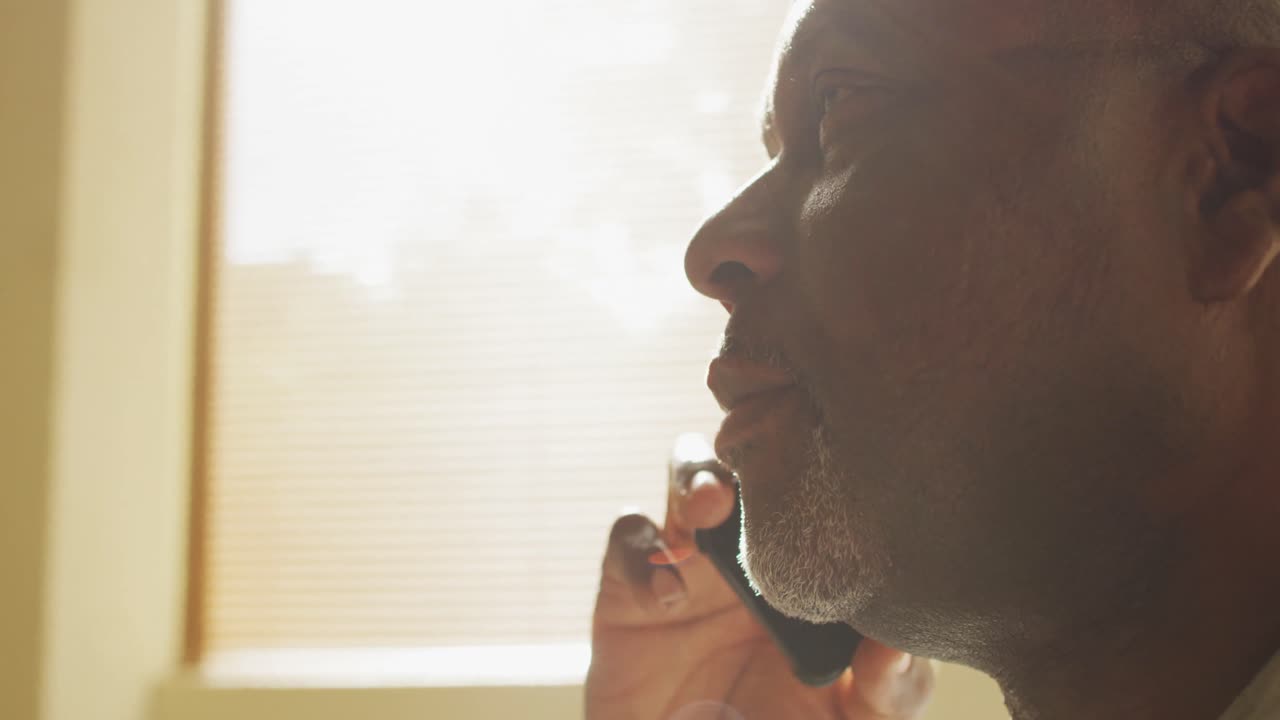 Thoughtful african american senior man sitting at home in the sun talking on smartphone