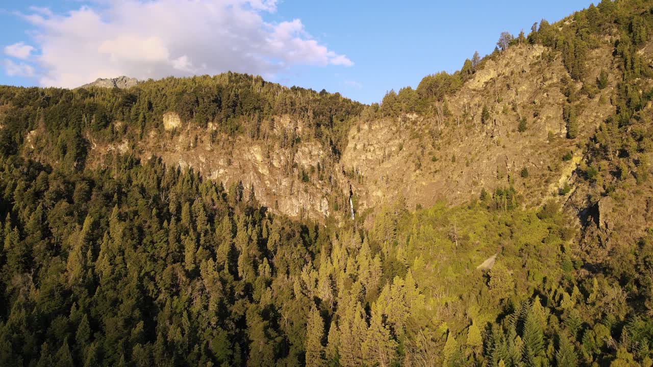 dolly aéreo en las montañas cubiertas de bosques de cipreses y cascada de corbata blanca en el fondo al atardecer, patagonia argentina