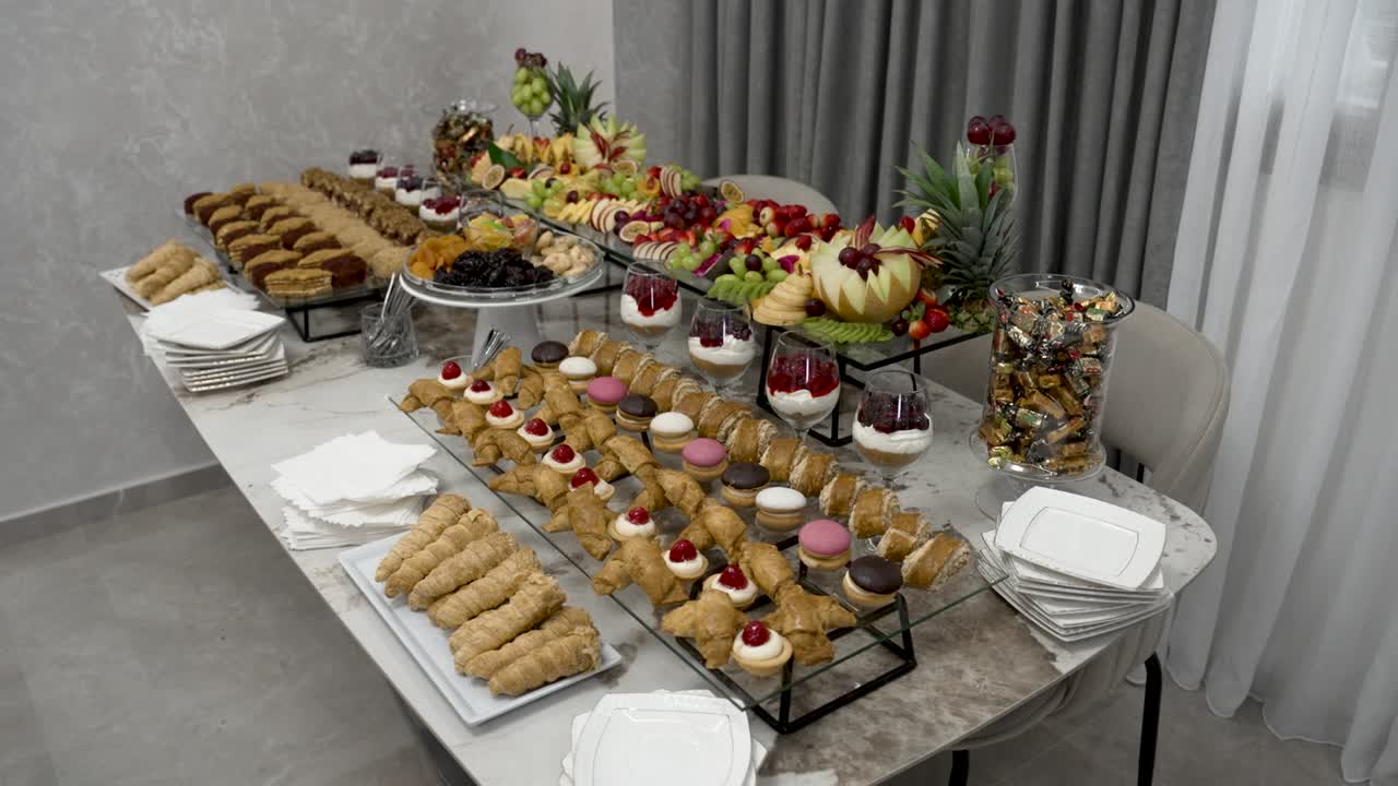 Un grande tavolo espone un assortimento di dolci e frutta a un evento di celebrazione. Diversi tipi di dolci sono disposti ordinatamente affinché gli ospiti possano gustarli. L'ambiente è luminoso e accogliente.