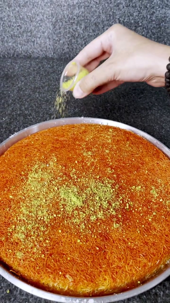 Close-up de uma mão polvilhando pistache finamente triturado sobre uma bandeja fresca de kunafa, uma sobremesa tradicional do Oriente Médio, em uma bancada escura de cozinha.