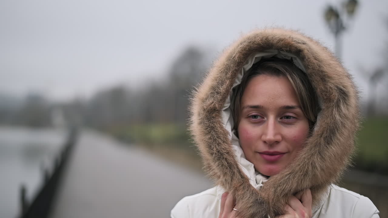 A woman smiles while walking along a path by the water on a cloudy day. She wears a warm coat with a fur hood, feeling comfortable in the cool weather