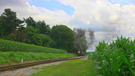 una vista frontal de un tren de pasajeros de vapor antiguo saliendo de una parada y viajando a través de tierras de cultivo y campos de maíz que soplan humo negro en un día soleado de verano