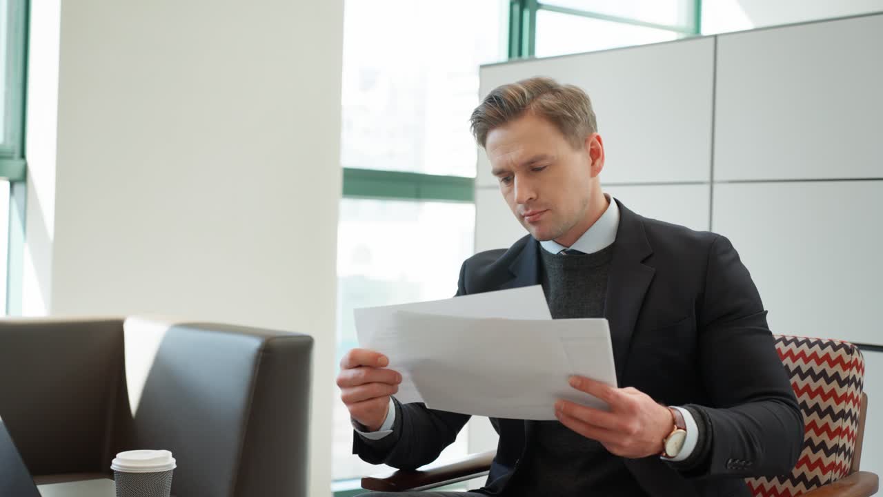 Blond Young professional man reviews business documents with diagrams and graphics while sitting in modern office space with large windows, laptop on the desk - dolly pull back