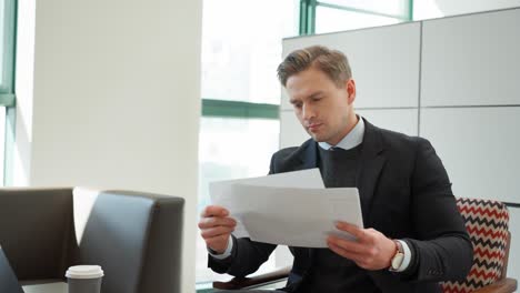 Blond Young professional man reviews business documents with diagrams and graphics while sitting in modern office space with large windows, laptop on the desk - dolly pull back