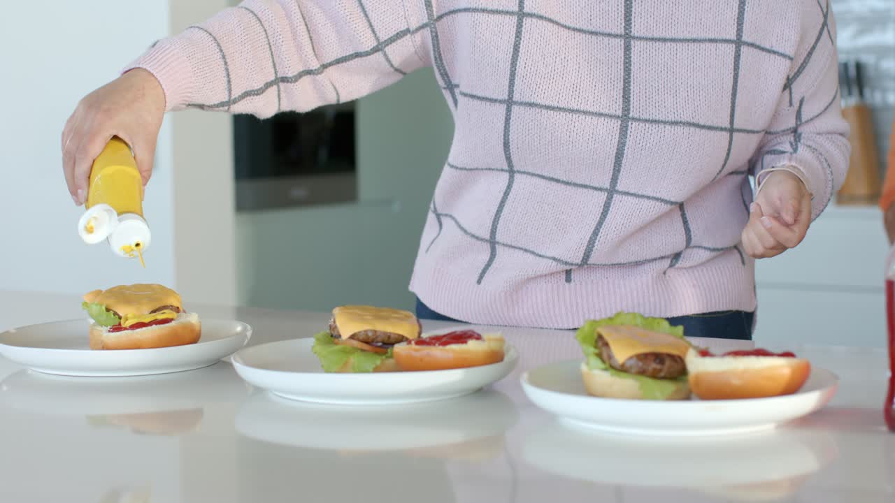 Reaching for yellow bottle, friends squeezing mustard onto buns at counter while man stepping in