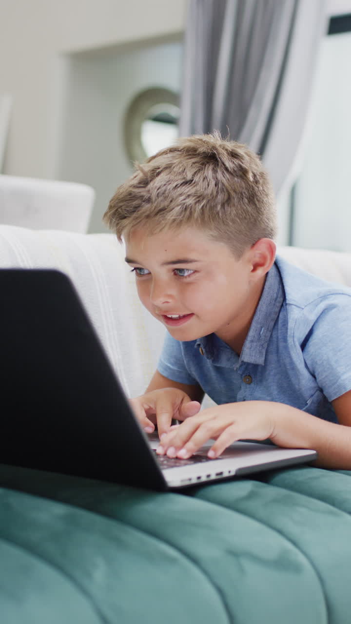 Vertical video of happy caucasian boy lying on sofa and using laptop, slow motion