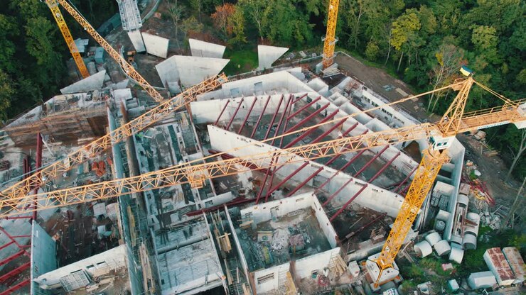 Luftaufnahme: Bau eines neuen Gebäudes mit hohen Turmkränen in grüner Umgebung.