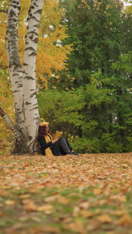 distant view of woman with red wavy hair wearing yellow beret and sunglasses flipping through book in her hand, face slightly blurred in background, surrounded by autumn trees, fallen leaves