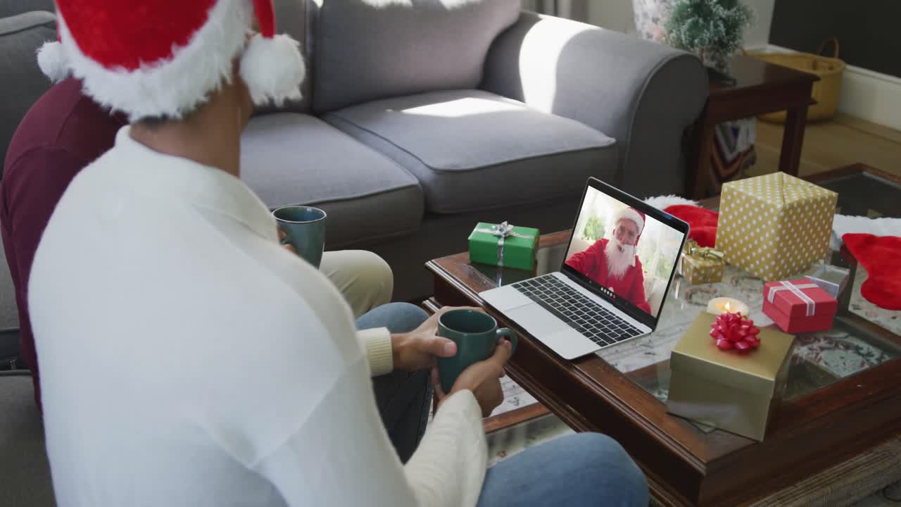 padre y hijo biraciales con sombreros de santa usando una computadora portátil para una videollamada de navidad con santa en la pantalla
