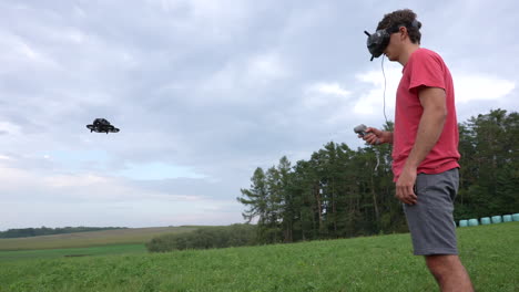 hombre con gafas, auriculares y controlador de movimiento parado frente a un dron fpv en una pradera