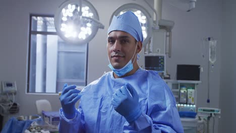 Portrait-of-mixed-race-male-surgeon-wearing-lowered-face-mask-smiling-in-operating-theatre