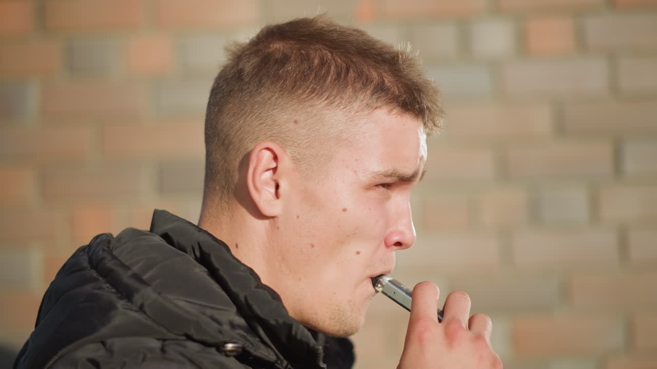 side view young man using vaping device puffing smoke under bright sunlight with soft shadows and blurred brick wall background in urban area