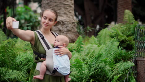 a young woman travels with her baby in a kangaroo backpack, snapping pictures on a mobile phone. while walking, she occasionally checks the phone screen