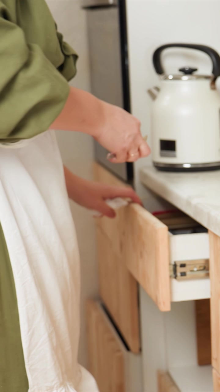 Woman in green top and white apron cleans kitchen surfaces. Opens wooden drawer to organize items inside closes cabinet doors. Kitchen setting includes countertop appliances