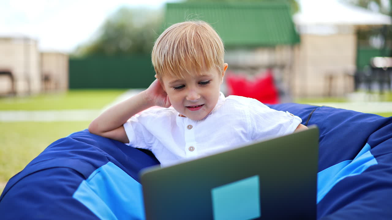 Lovely blond kid using gadgets outdoors. Nice little boy sitting in bean bag chair watching videos on laptop smiling. Close up. Blurred backdrop.