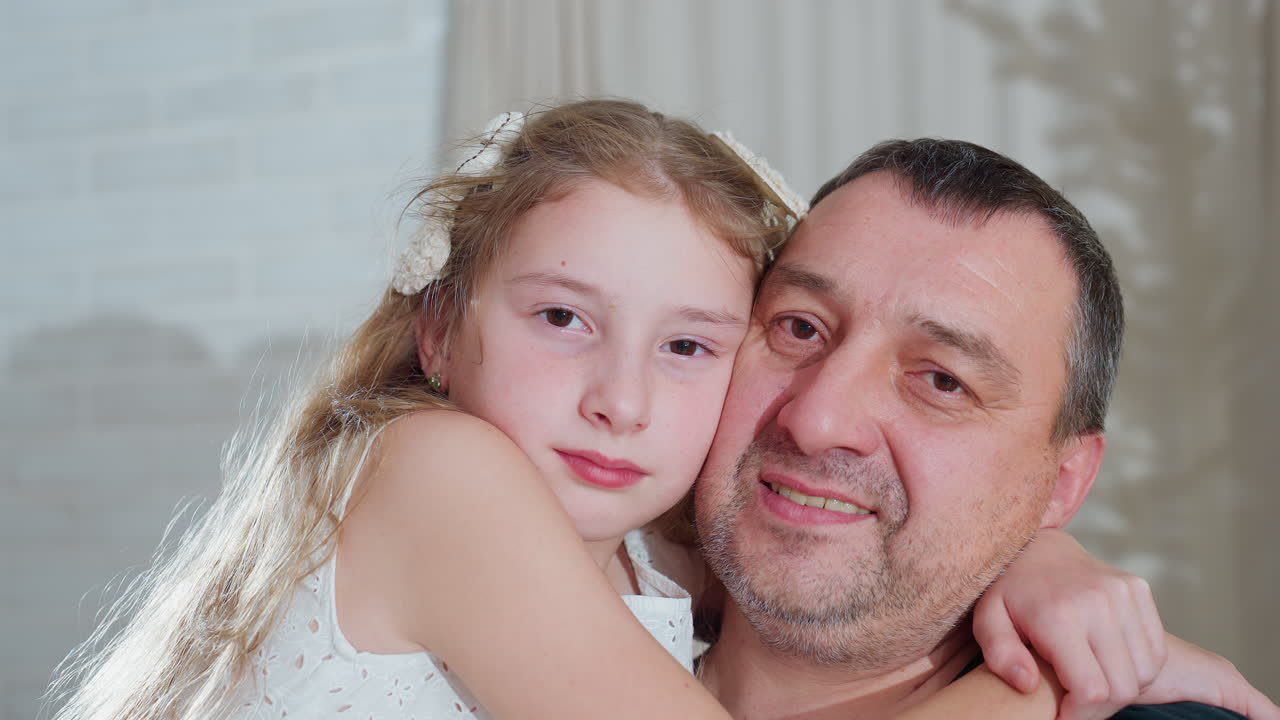 Heartwarming portrait view of young daughter in white gown embracing dad warmly around neck as they smile softly together capturing tender emotional family bond during cozy festive holiday season