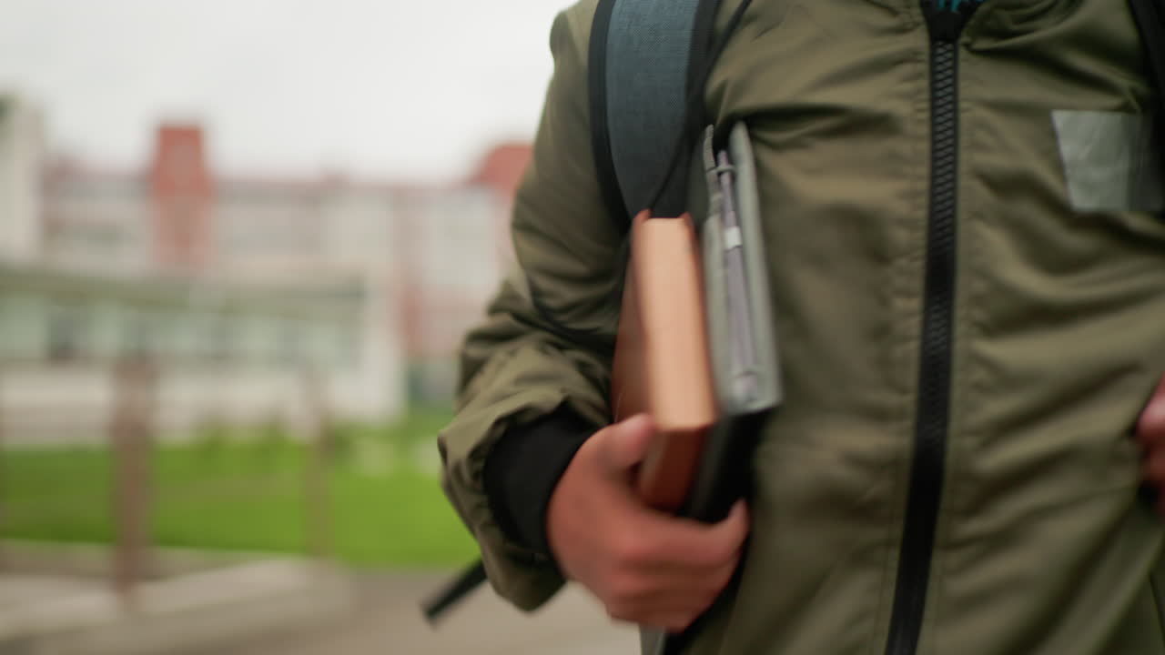 따뜻한 미소를 지으며 노트를 팔에 끼고 백팩을 멘 학생이 흐릿하게 보이는 대학 건물과 녹색 배경을 뒤로하고 달리는 모습이 클로즈업되어 쾌활한 학업 라이프스타일을 보여주고 있어요