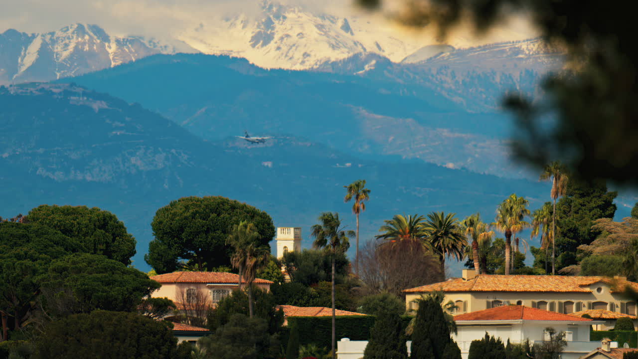 White airplane flying above buildings surrounded by green trees with the mountains on the background on a cloudy day