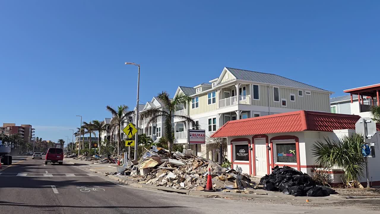A street in Indian Shores, Florida, lined with debris and damaged materials after Hurricane Milton, with palm trees and buildings under a clear blue sky.