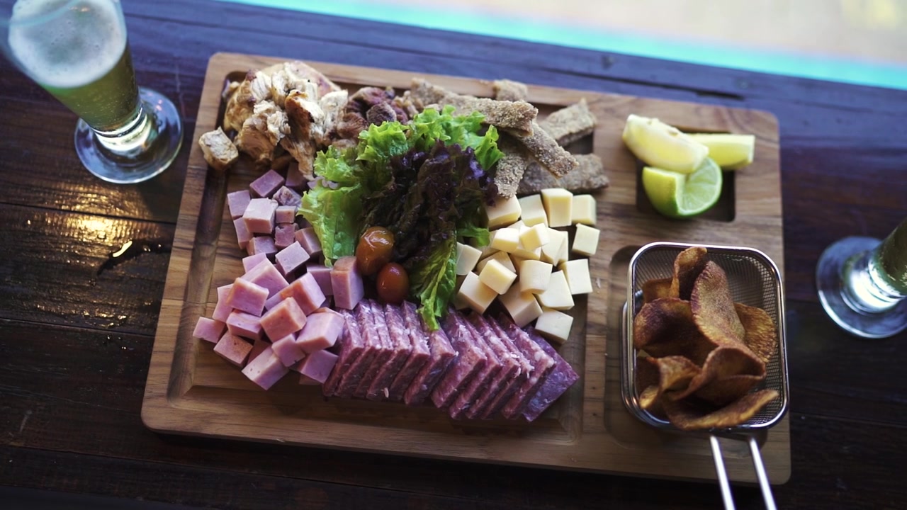 Hitaasti liikkuva ylhäältä päin otettu kuva ylellisestä charcuterie-tarjottimesta, joka on asetettu ulkohotellibaarissa uima-altaan vieressä. Tyylikäs esillepano ja rauhallinen uima-altaan ympäristö korostavat ylellistä tunnelmaa.