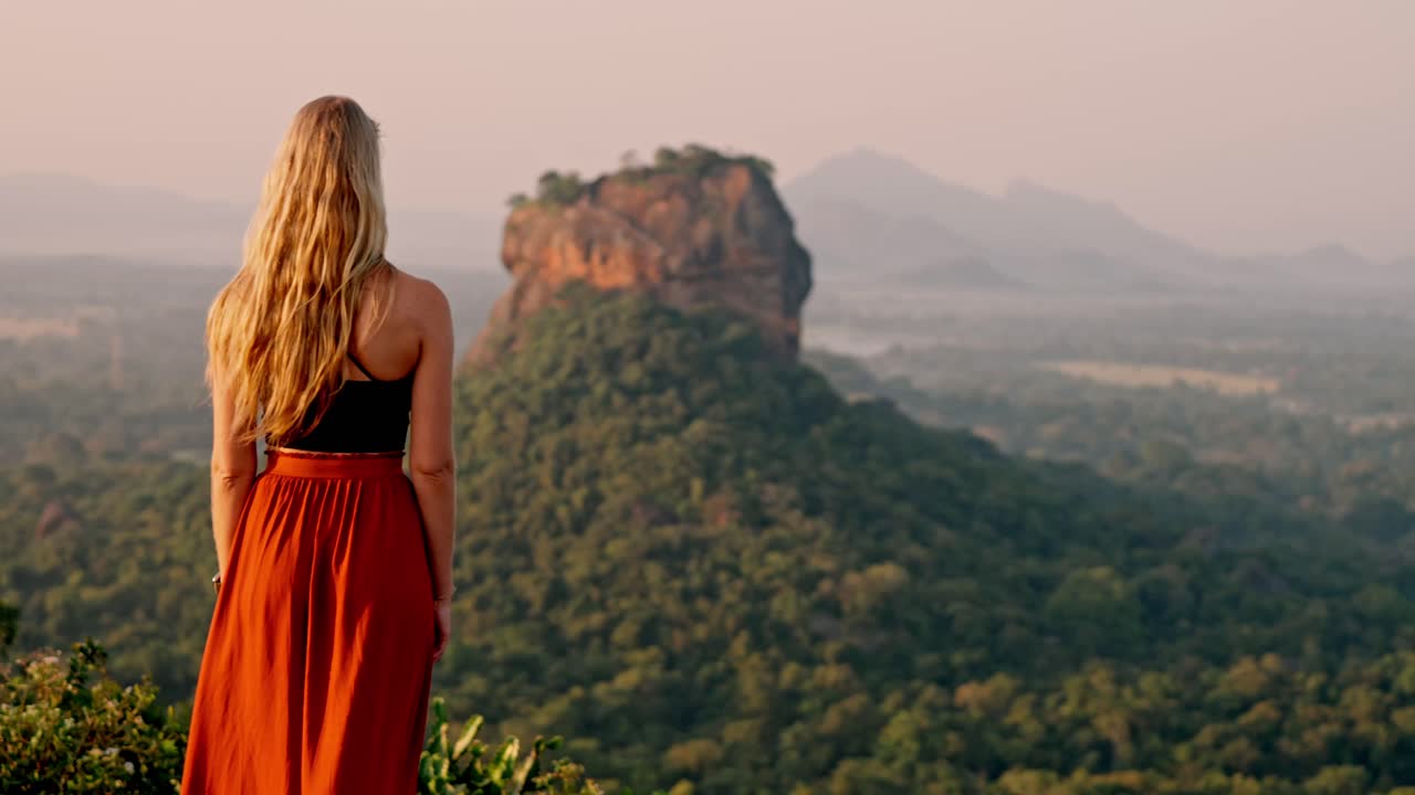 A lone woman stands amid lush greenery, gazing toward the ancient Sigiriya Rock fortress as the sun rises over Sri Lanka.