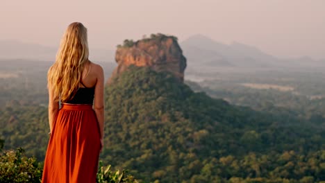 a lone woman stands amid lush greenery, gazing toward the ancient sigiriya rock fortress as the sun rises over sri lanka.