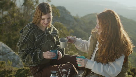 dos jóvenes excursionistas bebiendo té caliente del termo y hablando mientras se sientan en el acantilado de la montaña en un soleado día de otoño