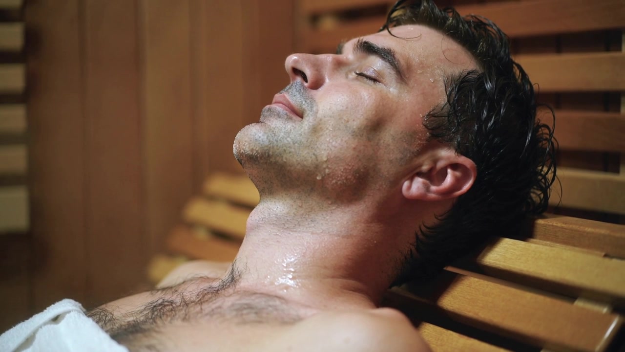 Uomo sdraiato che respira su una panca di legno in sauna, con un asciugamano bianco sul petto e il sudore che brilla mentre si riposa.