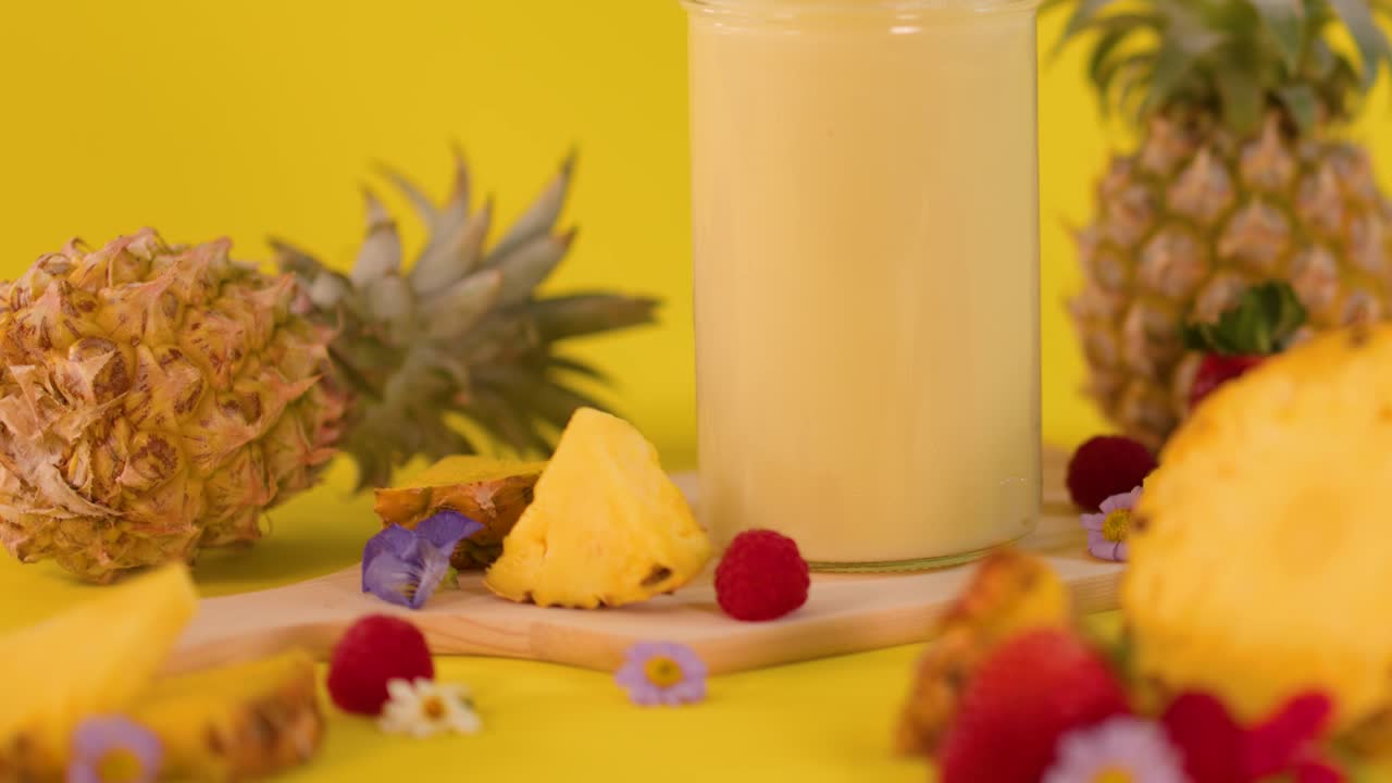 Hand adds whipped cream to pineapple smoothie with fresh fruit, bright yellow background, even lighting