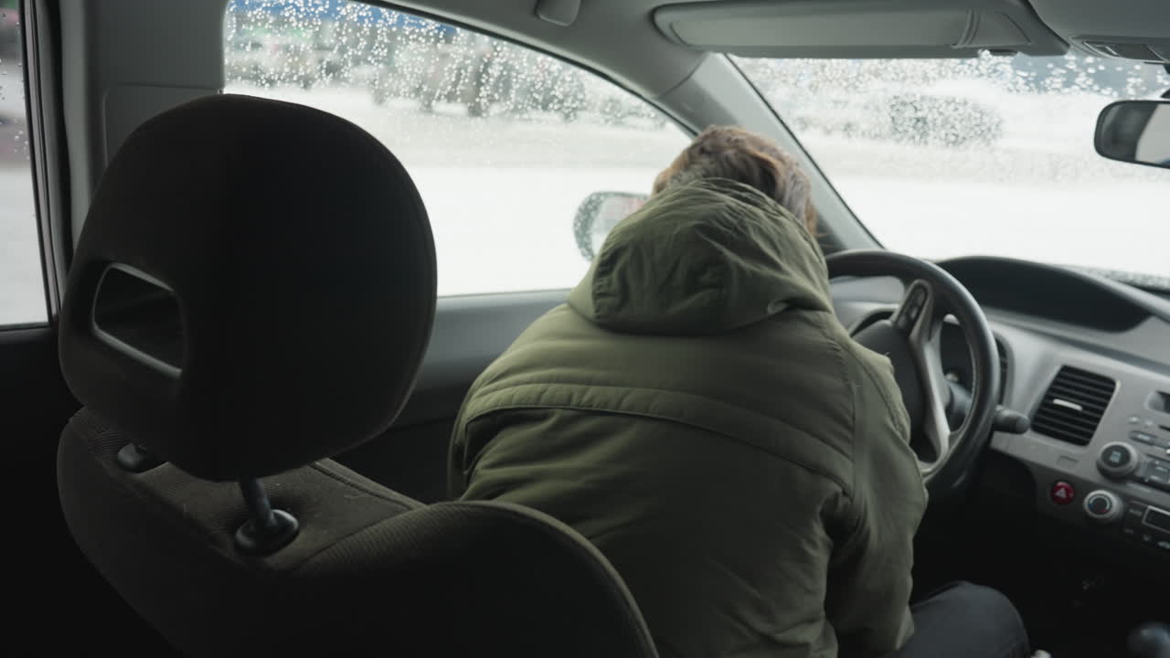 young man in winter jacket seated inside vehicle closes door and adjusts driver seat while parked cars blur through window with raindrops during overcast cold weather