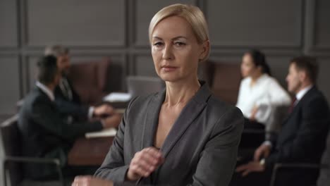 portrait of a serious businesswoman standing with crossed arms and looking at camera while in blurred background group of business executives sitting around table during meeting