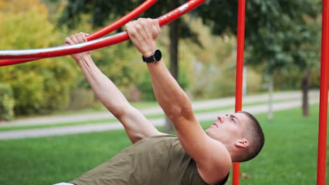 Focused young muscular athletic man doing pull-ups at red sports facility in park