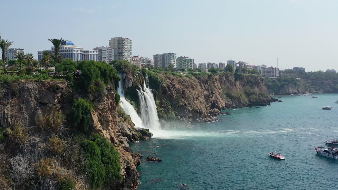 Drone aereo che sorvola una scogliera rivelando le acque delle Cascate del Duden Inferiore che precipitano da una scogliera rocciosa direttamente nel Mar Mediterraneo ad Antalya, Turchia, mentre le barche turistiche osservano dal basso