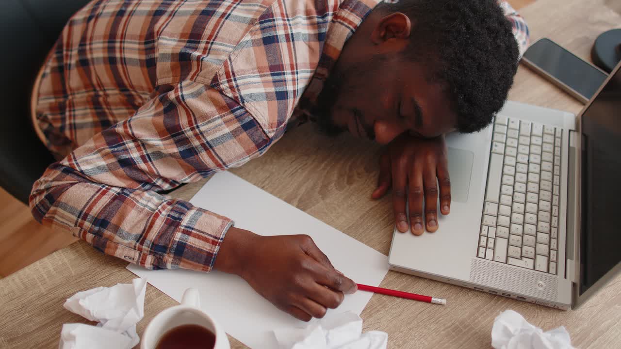 Tired man at home office falling asleep on table with laptop computer crumpled sheets of paper