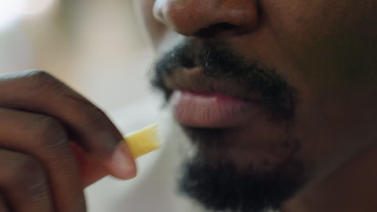 Primo piano di un uomo che addenta una patatina fritta, con le dita che toccano delicatamente le labbra durante un pasto al chiuso, che mostra baffi, barba, la grana della pelle ed espressione naturale in un ambiente con luce naturale soffusa