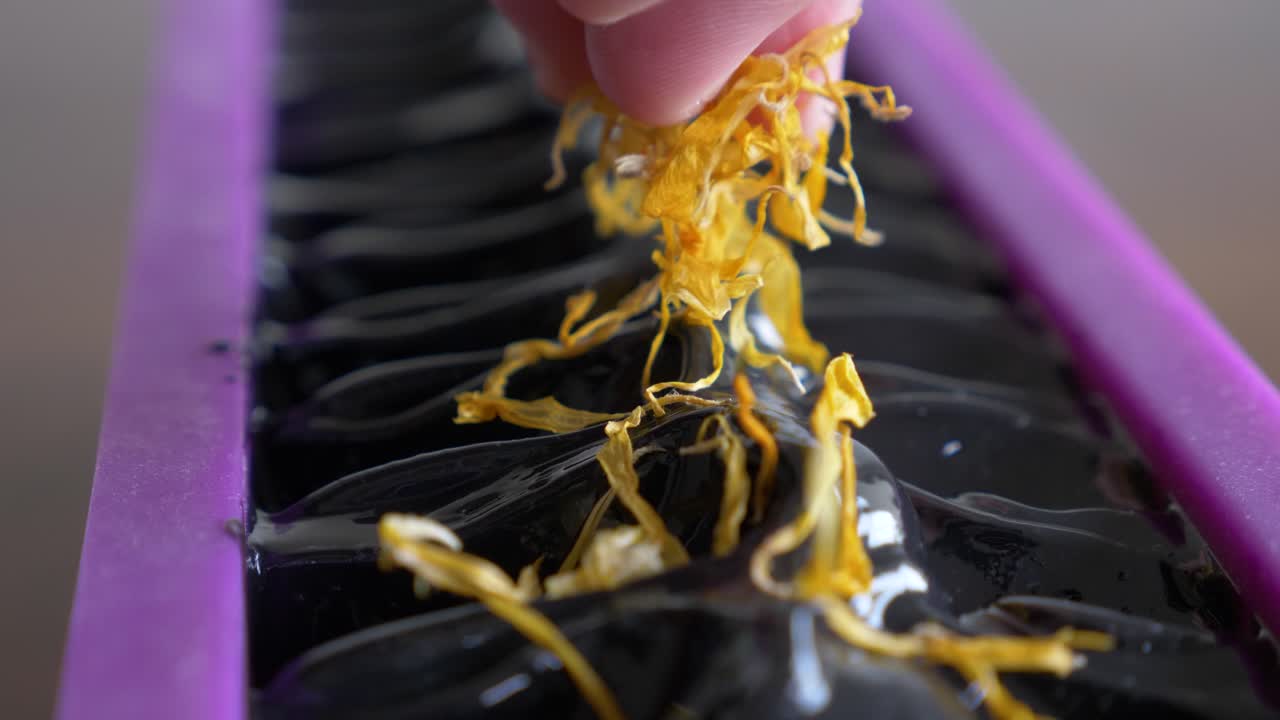 한 여성이 새로 만든 검은 비누 블록을 노란색 꽃의 말린 꽃잎을 뿌려 부드럽게 장식합니다.