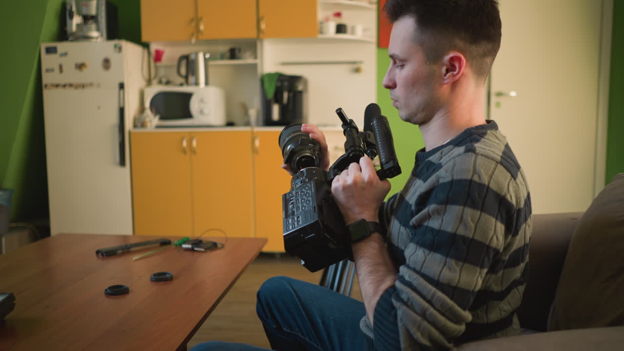 Person sitting on sofa in home studio, focusing on video camera setup with tripod, preparing for filming project. Workspace with coffee machine and colorful objects in background