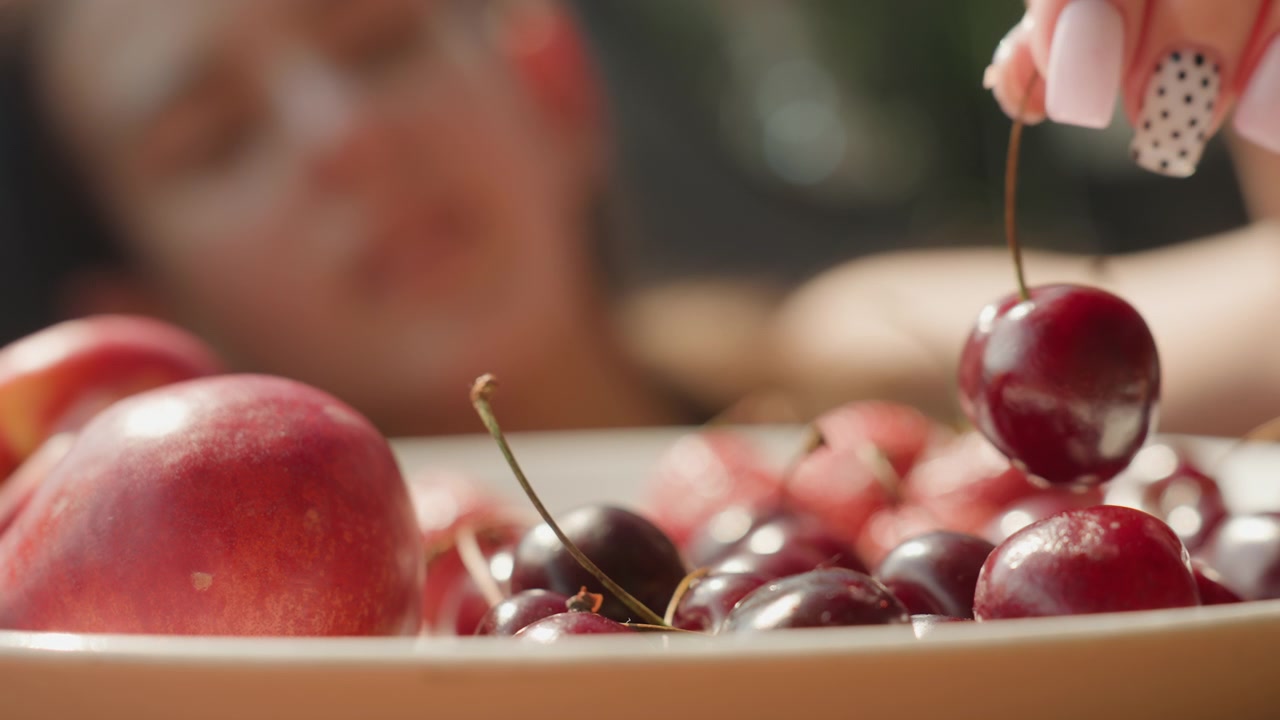 Primo piano di frutta fresca, tra cui ciliegie e pesca, disposta in una ciotola con illuminazione soffusa. Una persona è visibile fuori fuoco mentre raccoglie o si protende per i frutti. Scena.